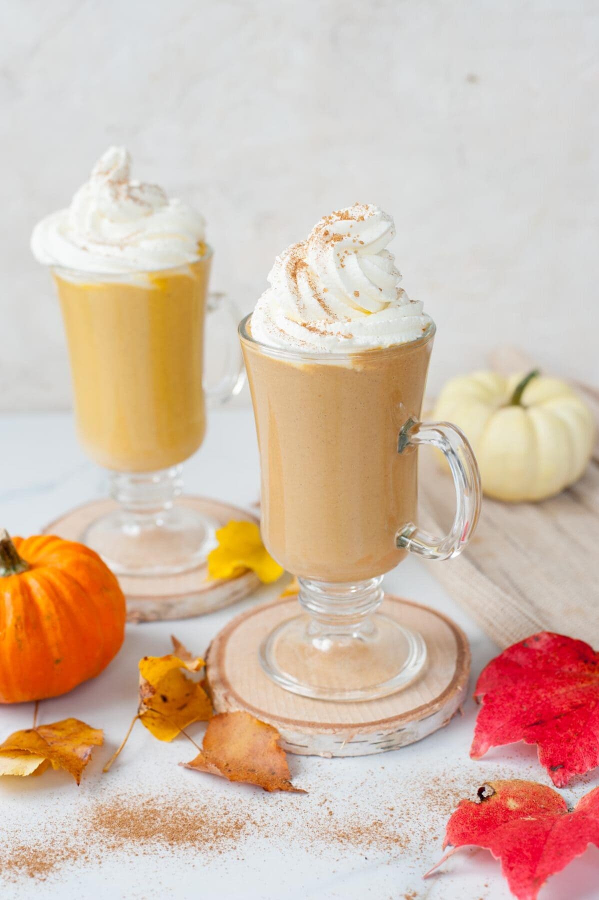 Two glasses with pumpkin hot chocolate topped with whipped cream. Small pumpkin and leaves scattered around.