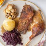 Roast goose leg and breast on a brown plate with braised cabbage and potato dumplings.