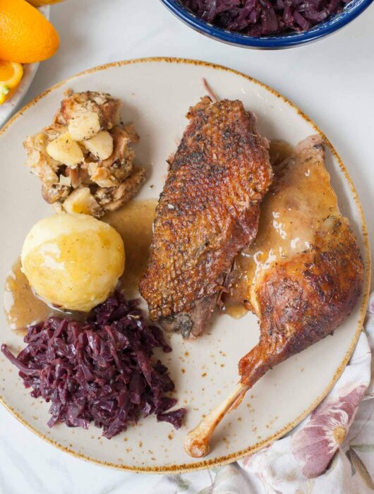 Roast goose leg and breast on a brown plate with braised cabbage and potato dumplings.