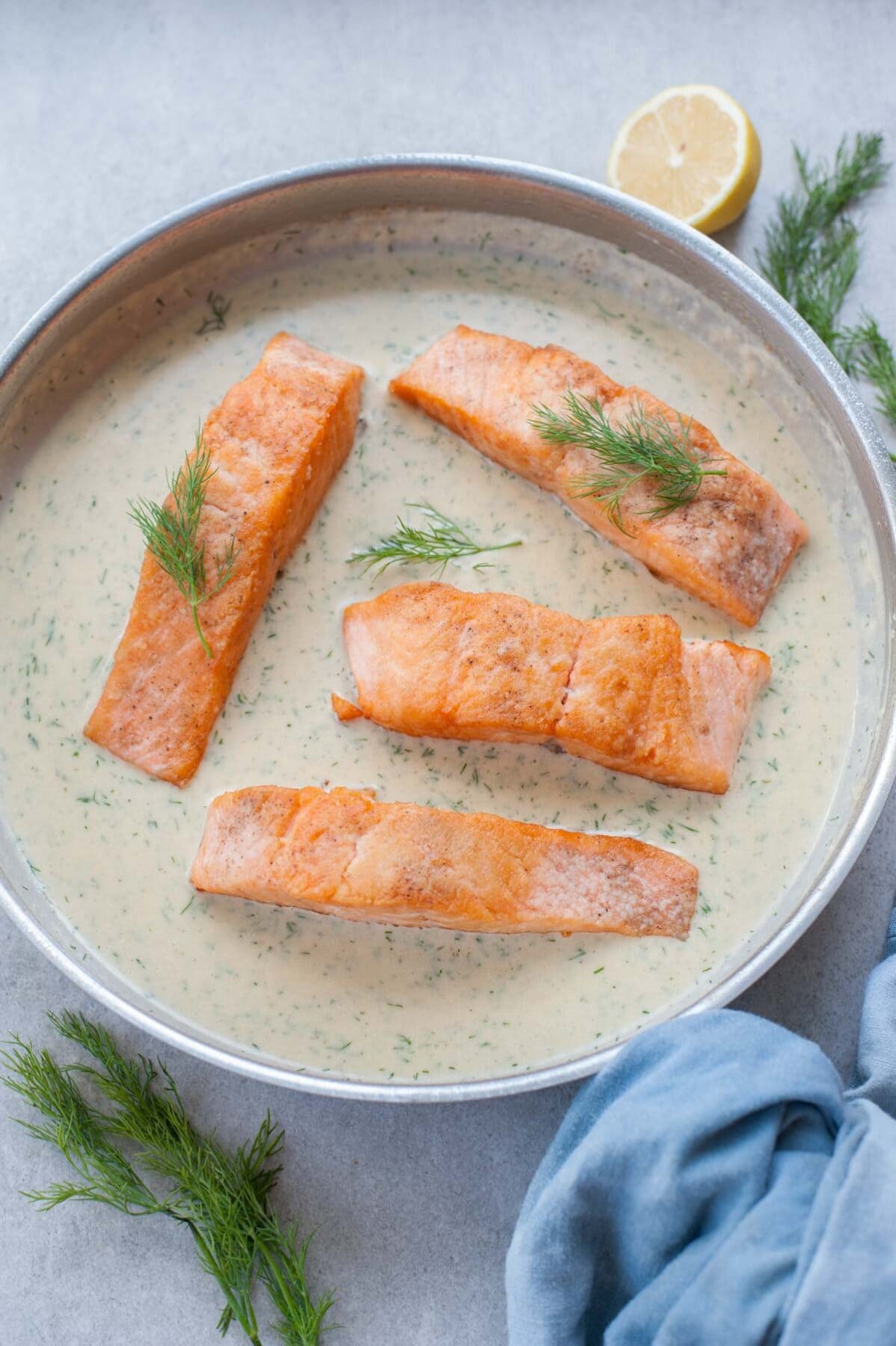 Salmon with creamy dill sauce in a frying pan. Blue kitchen cloth and dill in the background.
