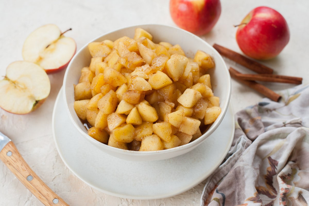 Sautéed Cinnamon Apples in a white bowl. Apples and cinnamon in the background.