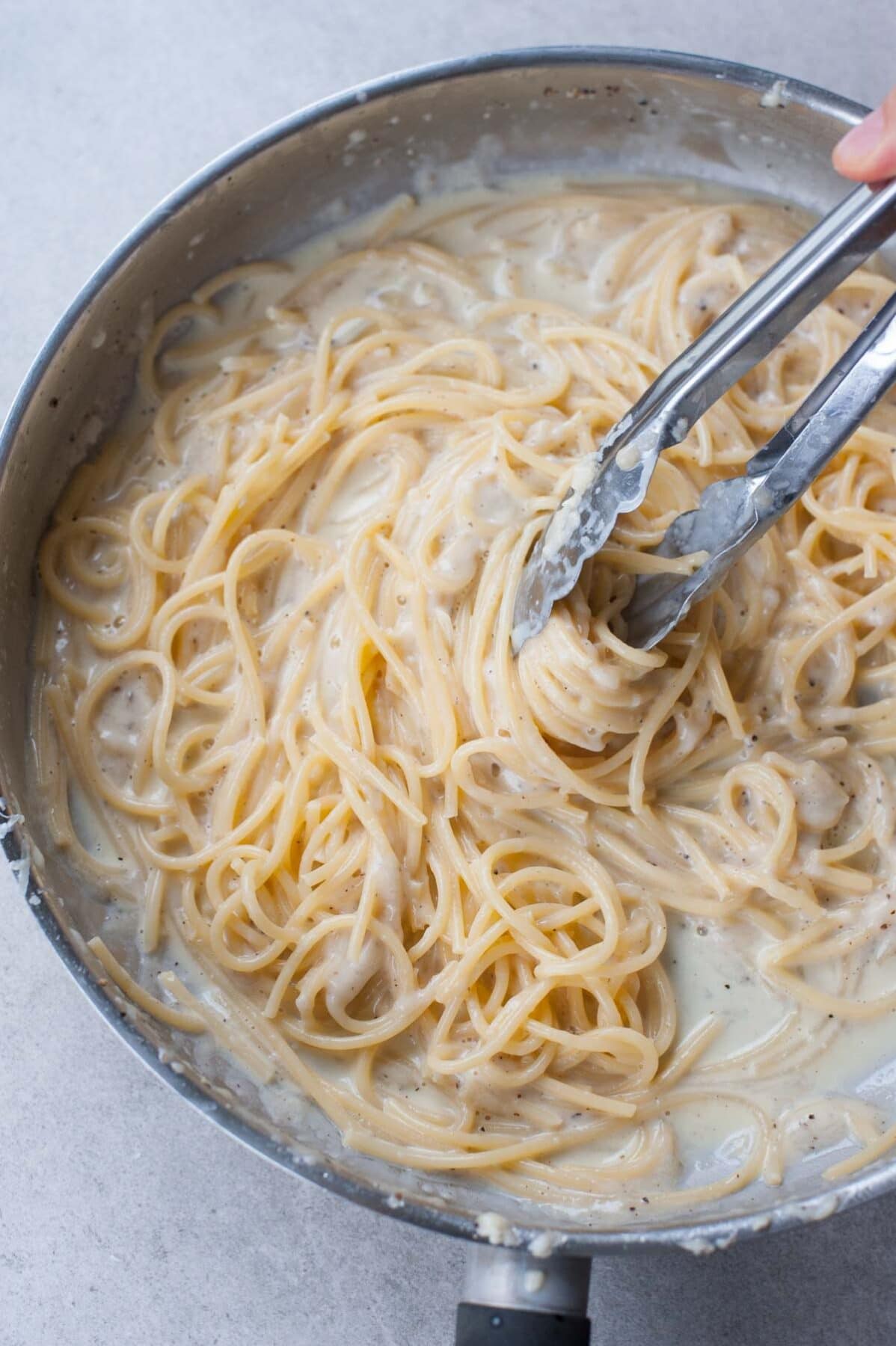 Spaghetti cacio e pepe in a metal pan stirred with kitchen tongs.