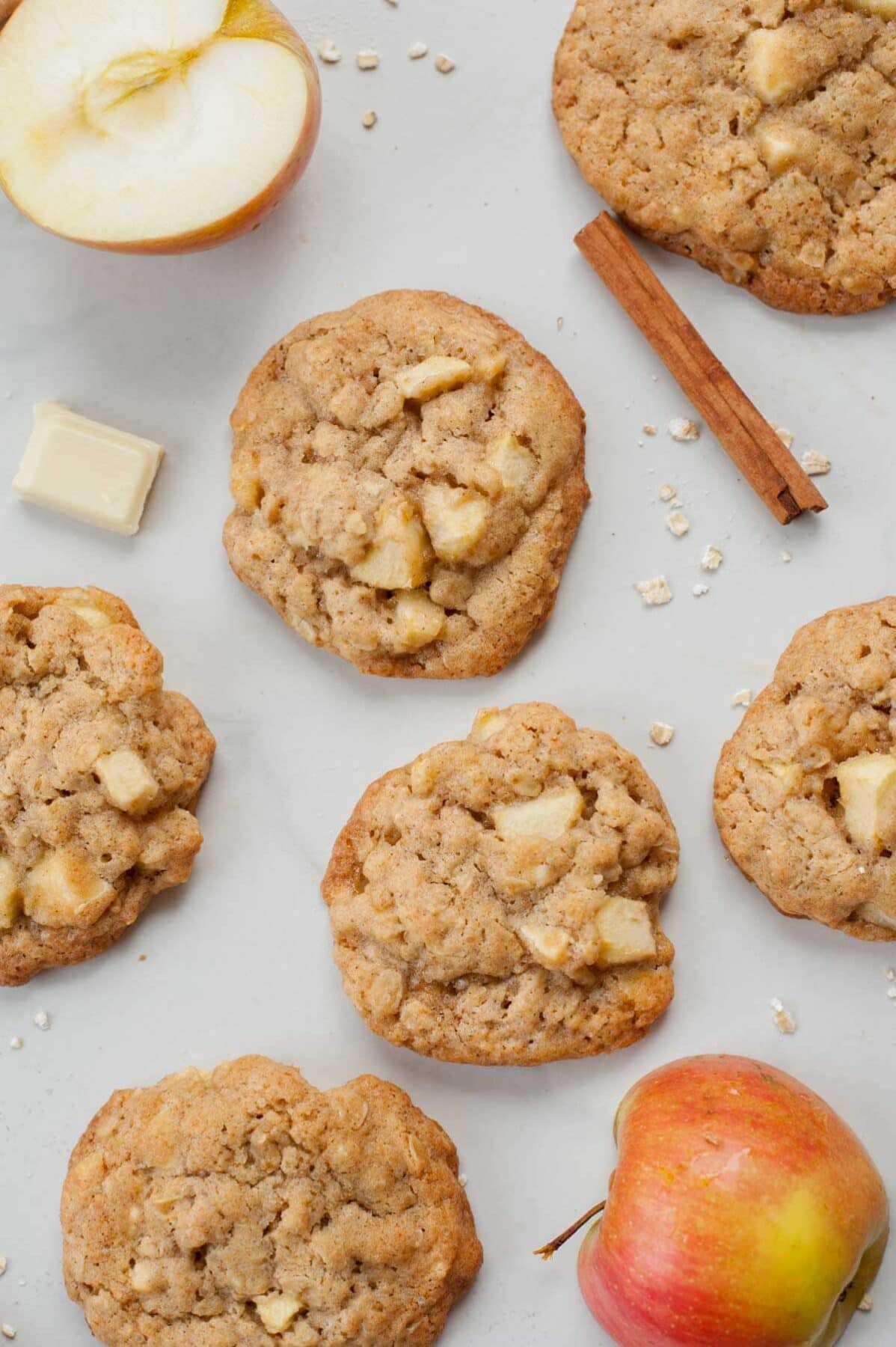 Apple cinnamon oatmeal cookies on a white table. Apple halves, white chocolate and oats scattered around.