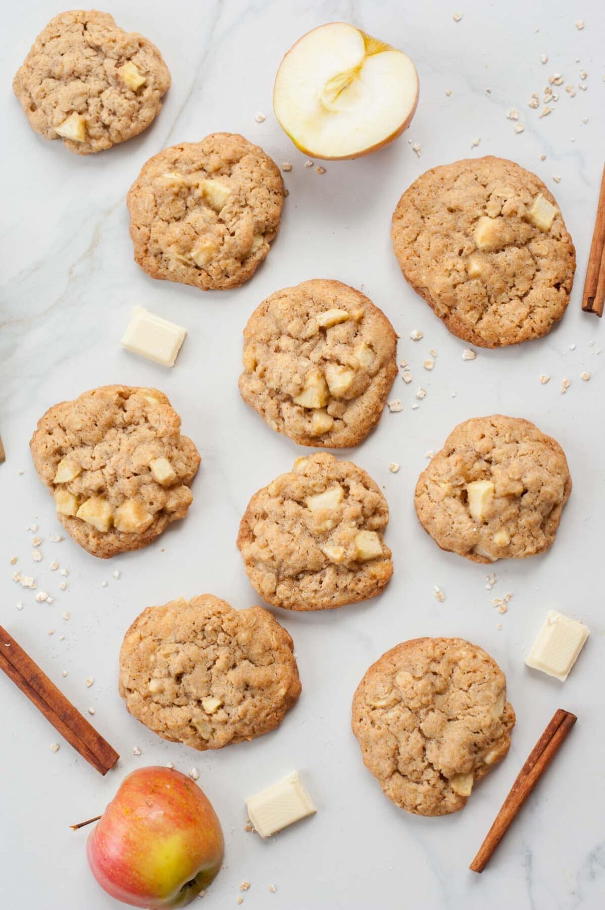 Apple cinnamon oatmeal cookies on a white table. Apple halves, white chocolate and oats scattered around.