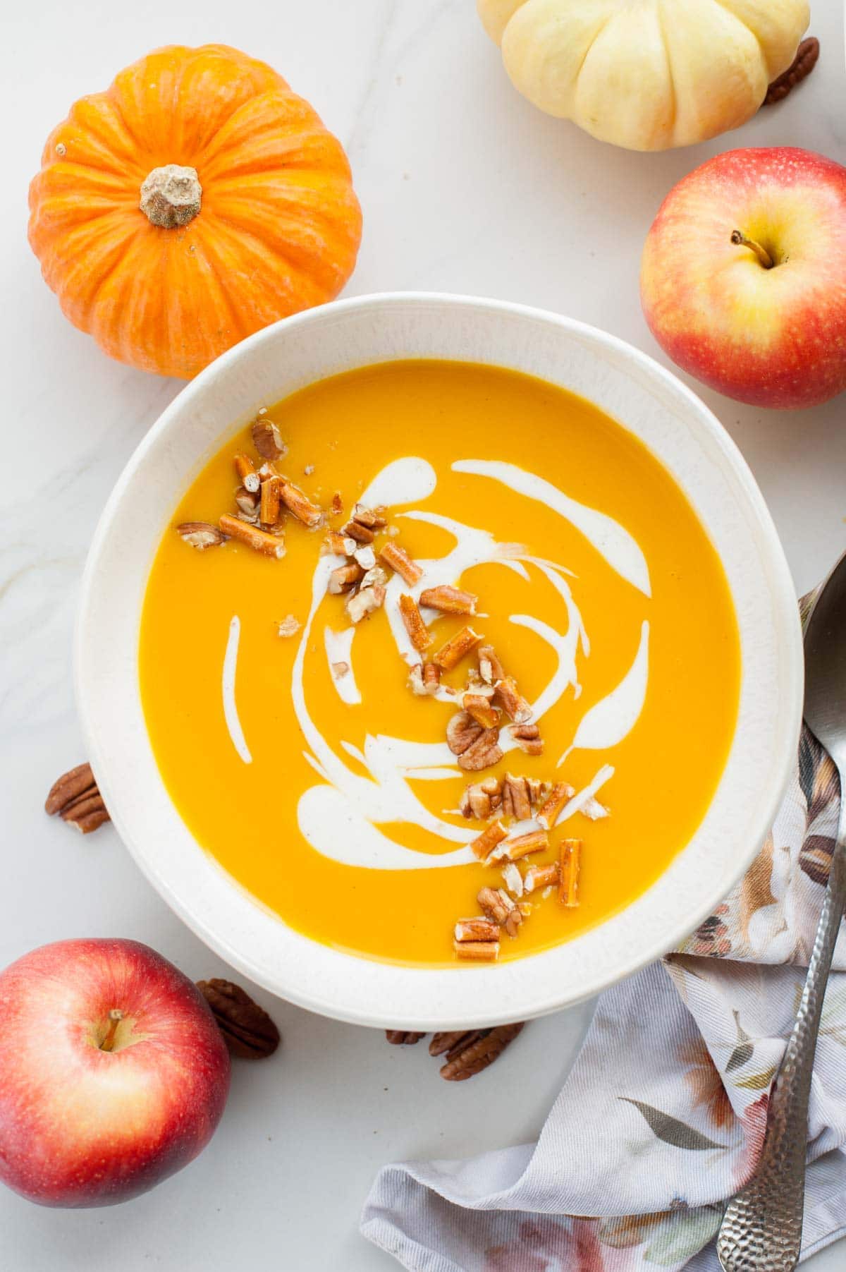 White bowl with butternut squash soup topped with yogurt and pecans. Apples in the bachground.