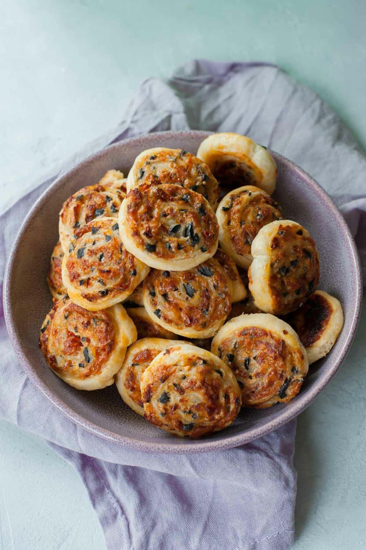 Cheese sun-dried tomato and olive puff pastry pinwheels in a violet bowl.