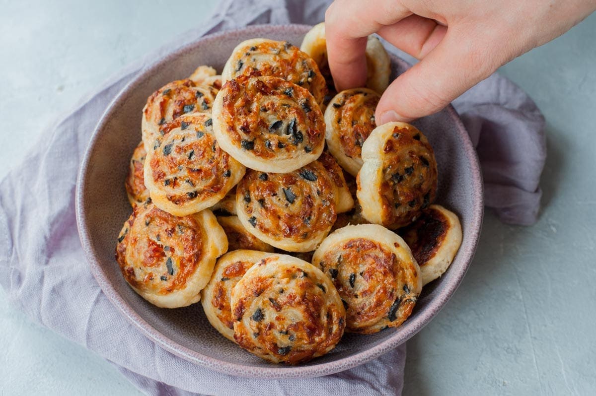 Cheese sun-dried tomato and olive puff pastry pinwheels are being picked up from a violet bowl.