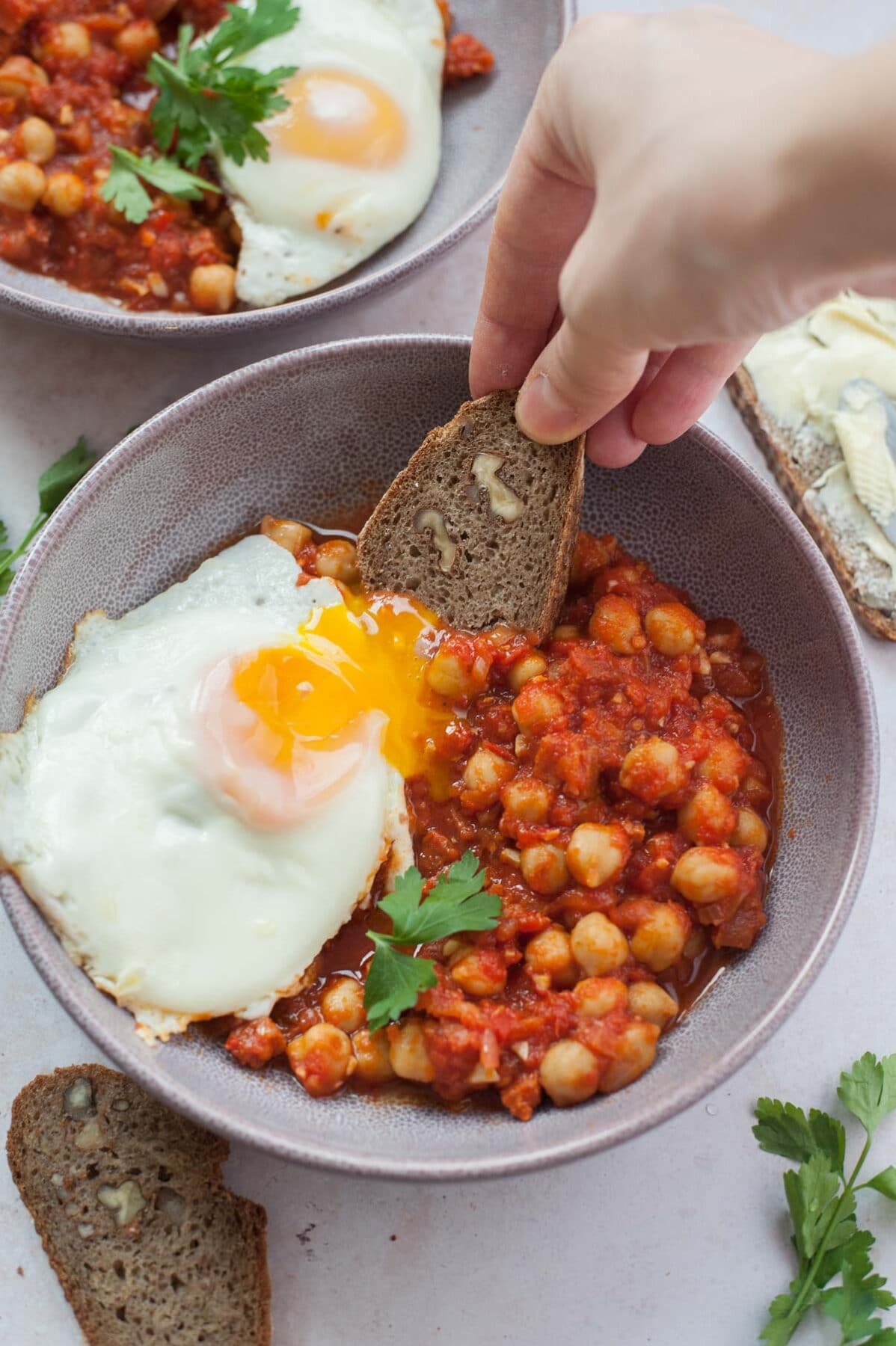 A piece of bread is being dunked in a runny egg yolk and tomato sauce with chickpeas and chorizo.