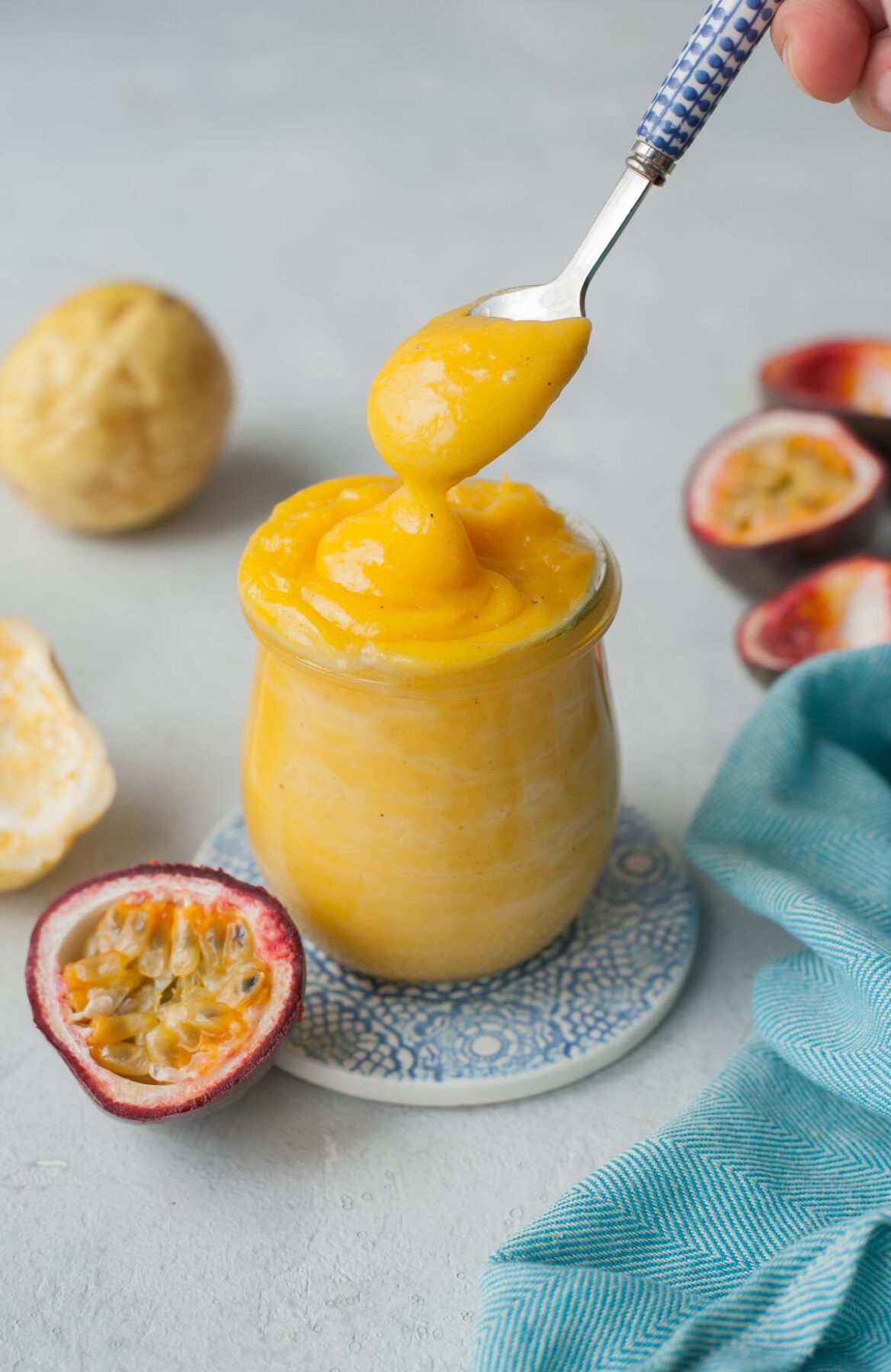 Passion fruit curd is being spooned into a jar. Passion fruits and a blue kitchen cloth in the background.