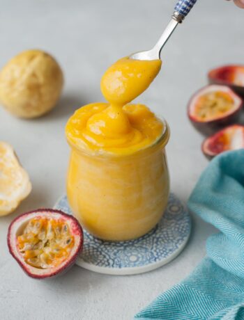 Passion fruit curd is being spooned into a jar. Passion fruits and a blue kitchen cloth in the background.
