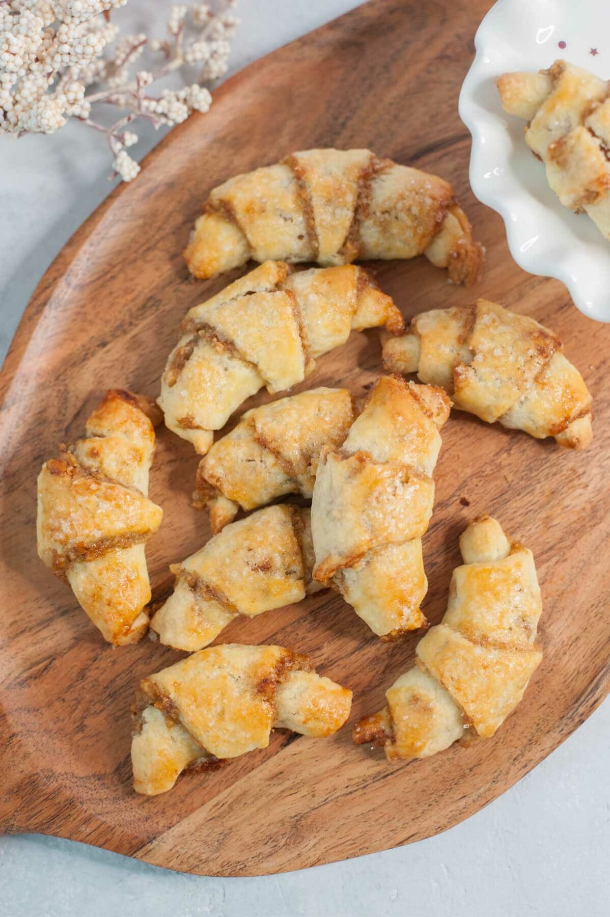 Orange walnut rugelach on a chopping board. White cake stand and white plants in the background.