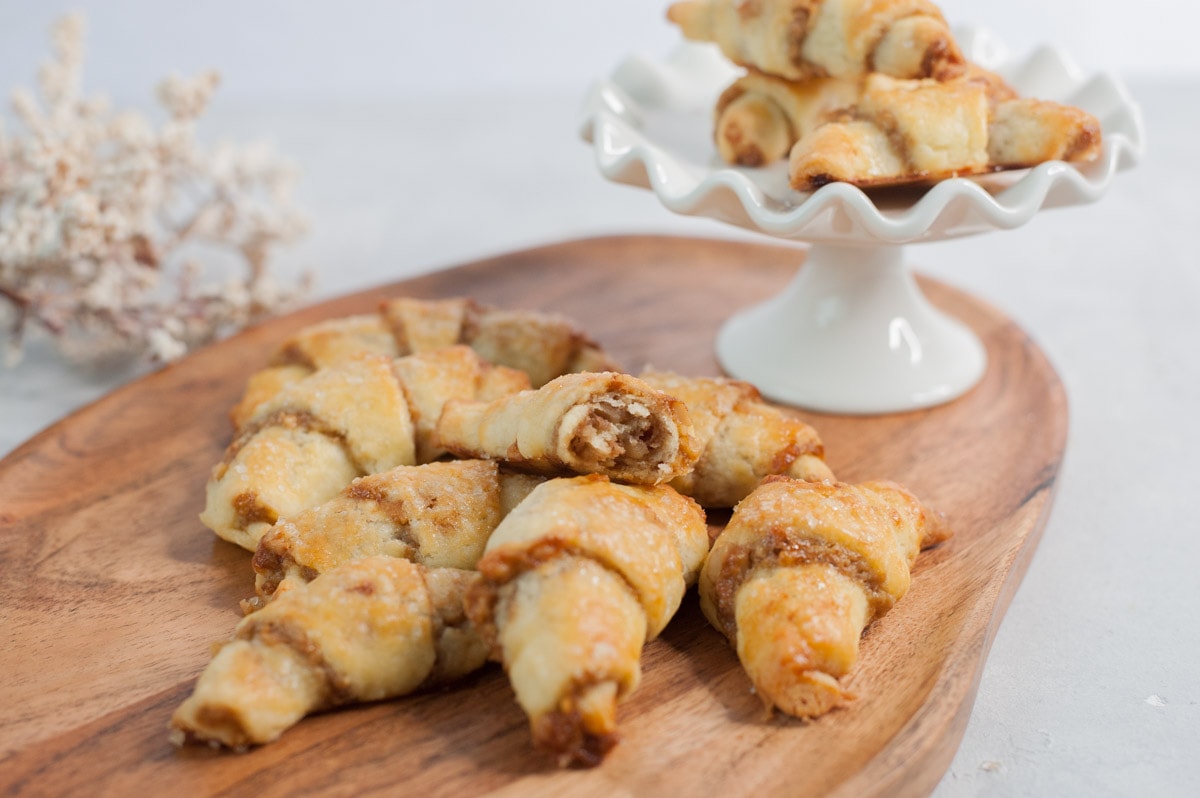 Orange walnut rugelach on a chopping board. White cake stand and white plants in the background.