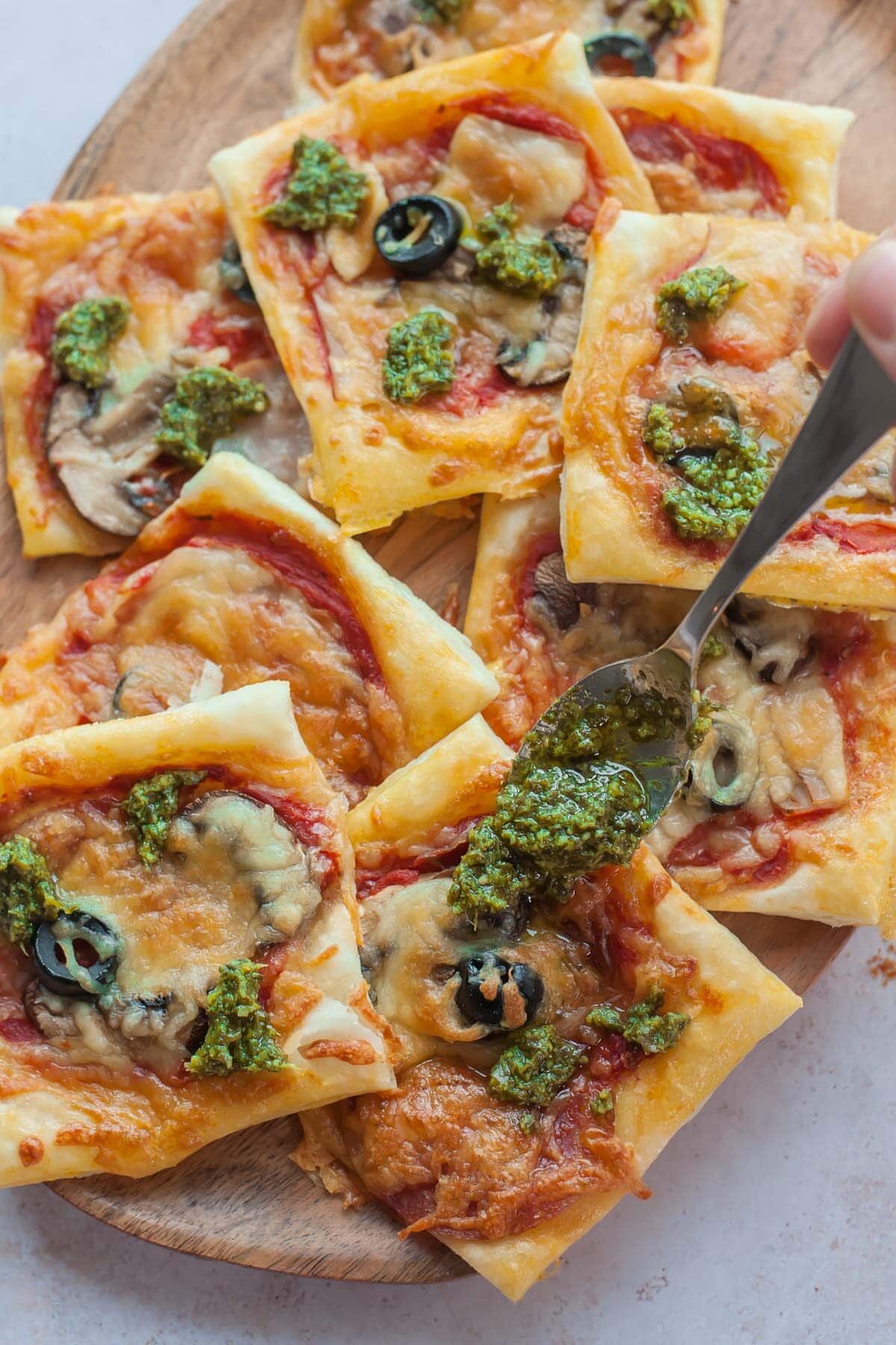 Pesto sauce is being spooned over puff pastry pizza bites.