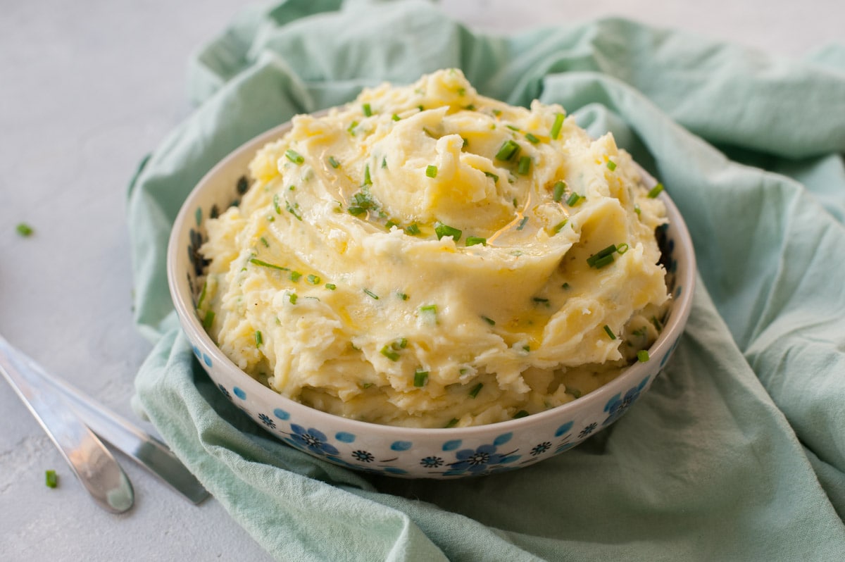 Sour cream mashed potatoes topped with melted butter and chives in a white-blue bowl. Green kitchen cloth in the background.