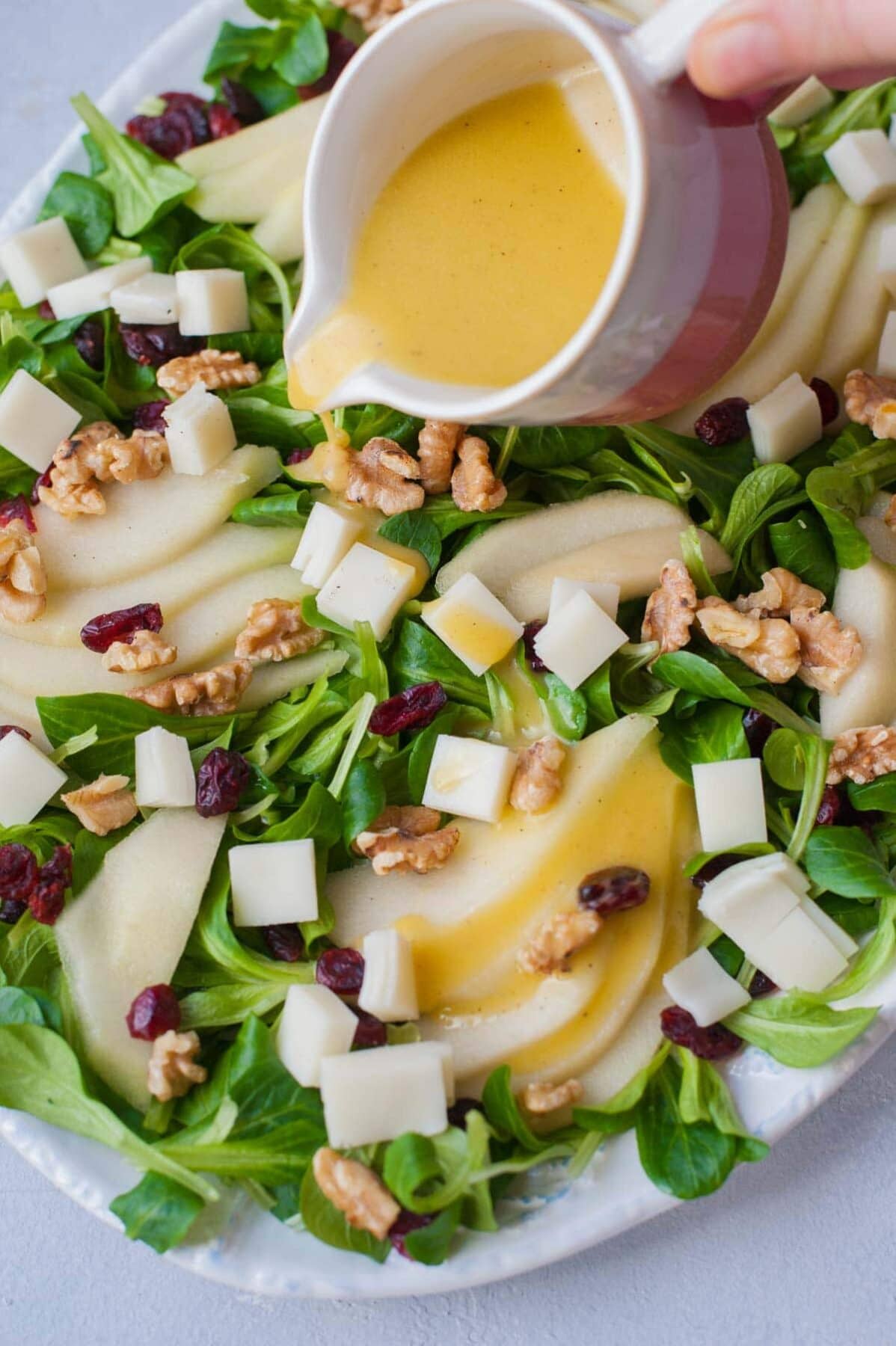 Lemon dijon vinaigrette is being poured over a pear walnut salad with cranberries and goat cheese.