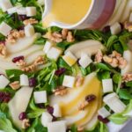 Lemon dijon vinaigrette is being poured over a pear walnut salad with cranberries and goat cheese.