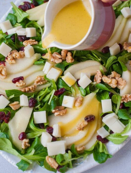 Lemon dijon vinaigrette is being poured over a pear walnut salad with cranberries and goat cheese.