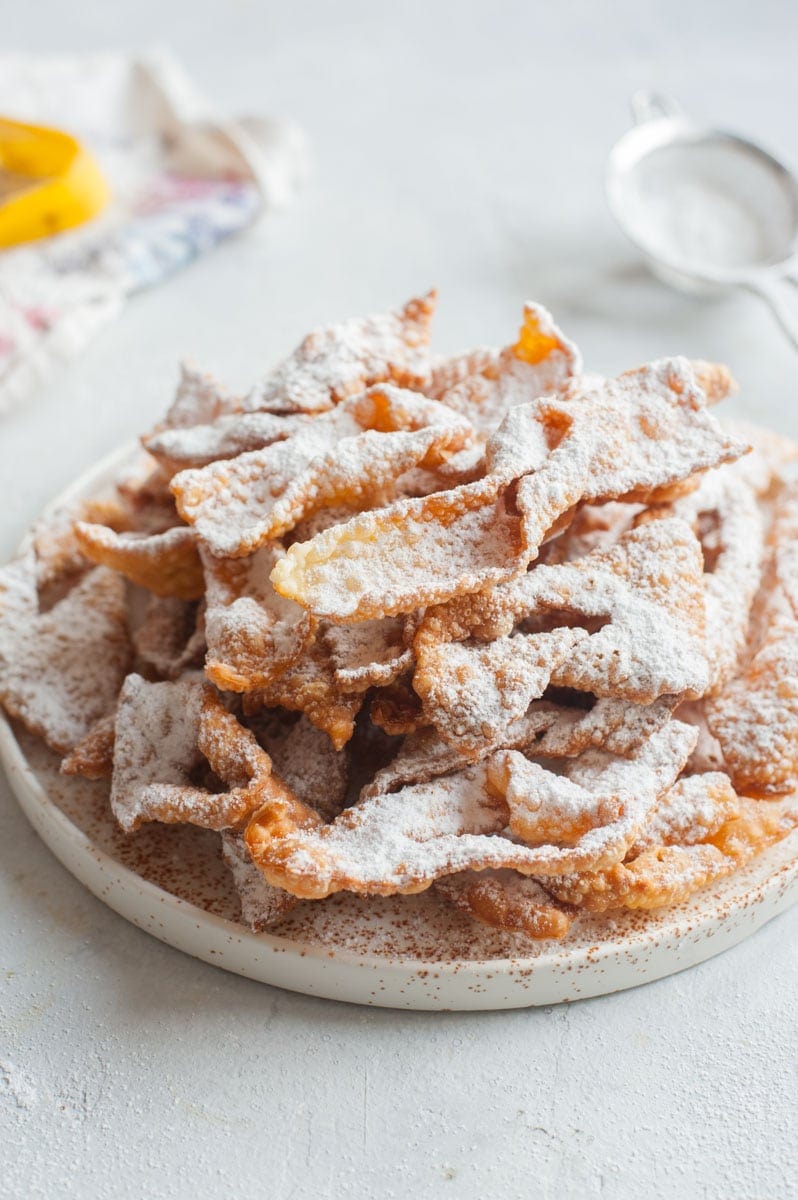 Faworki dusted with powdered sugar on a white plate. Kitchen cloth and a sieve in the background.