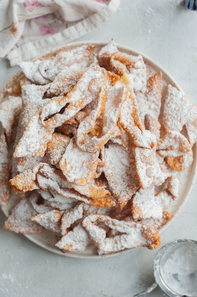 Faworki dusted with powdered sugar on a white plate. Kitchen cloth and a sieve in the background.