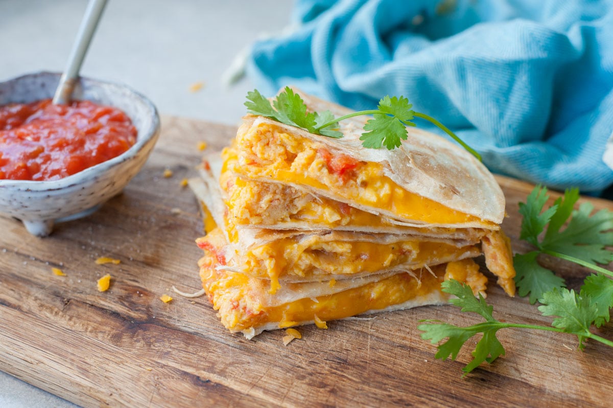 A stack of breakfast quesadillas on a wooden board. Tomato salsa and cilantro in the background.