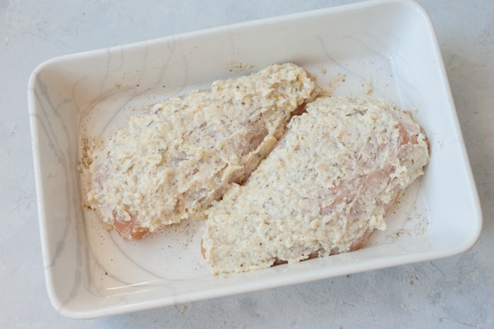 Chicken breasts in a white baking dish covered in mayo parmesan marinade.