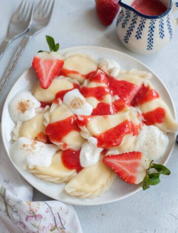 Sweet cheese pierogi on a white plate topped with strawberry sauce and whipped cream.