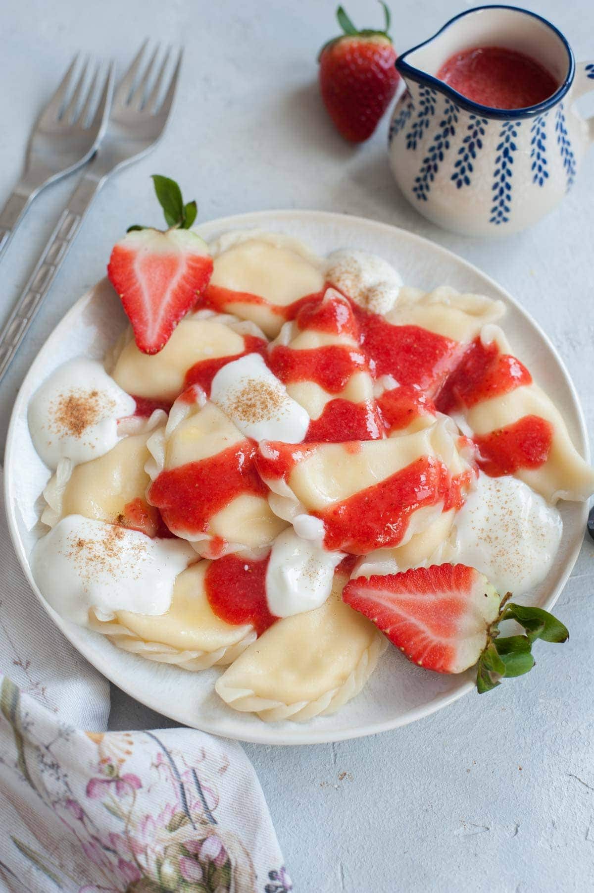 Sweet cheese pierogi on a white plate, topped with cream, strawberry sauce and fresh strawberries.
