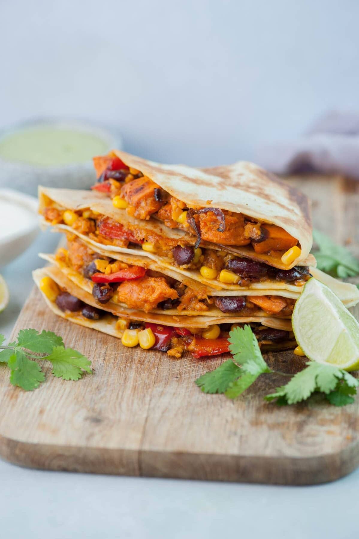 Sweet potato, beans, corn, and bell pepper quesadillas on a wooden chopping board, surrounded with cilantro leaves and limes.