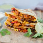 Vegetarian quesadillas on a wooden chopping board, surrounded with cilantro and lime quarters.