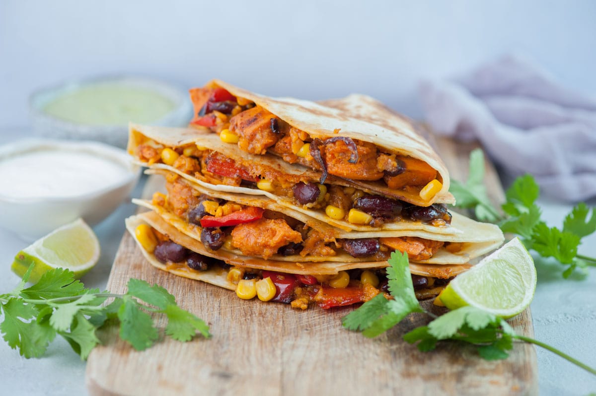 Sweet potato, beans, corn, and bell pepper quesadillas on a wooden chopping board, surrounded with cilantro leaves and limes.