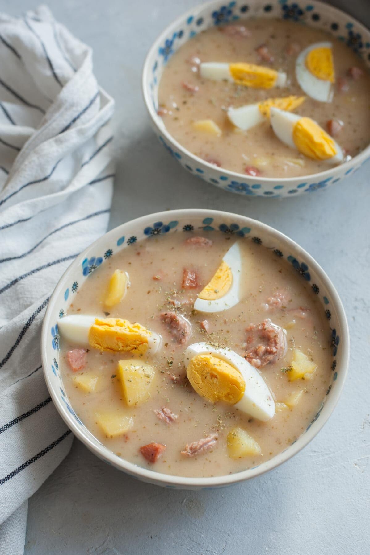 Two bowls with Żurek soup with kielbasa, eggs, and potatoes in a white-blue bowl.