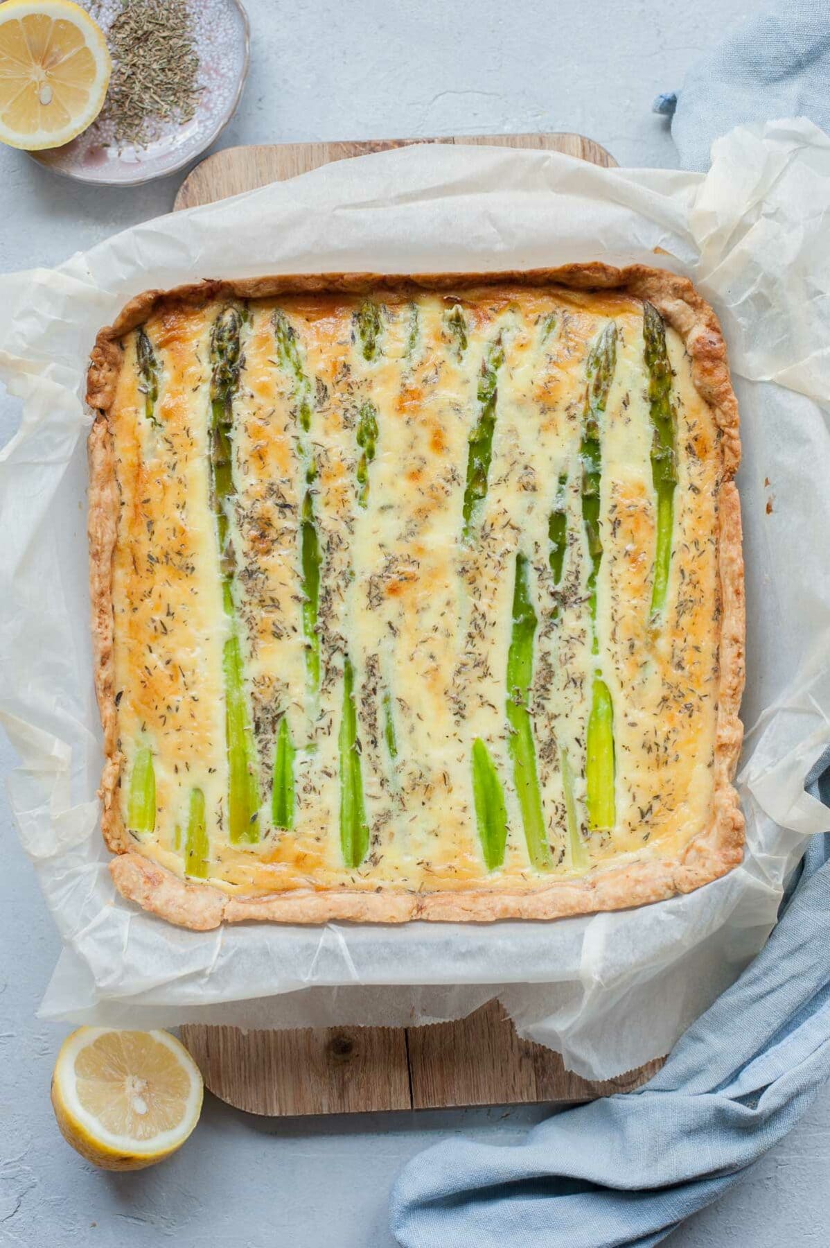 Asparagus Quiche on a wooden board. Kitchen towel and lemons in the background.