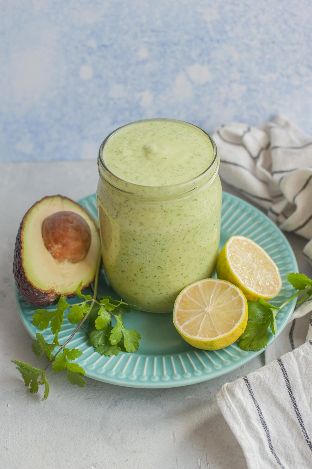 Avocado cilantro lime dressing in a jar on a green plate surrounded with avocado, limes and cilantro.