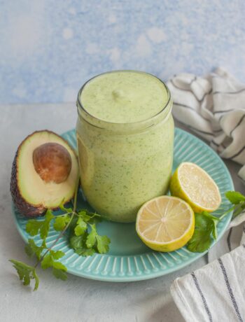 Avocado cilantro lime dressing in a jar on a green plate surrounded with avocado, limes and cilantro.