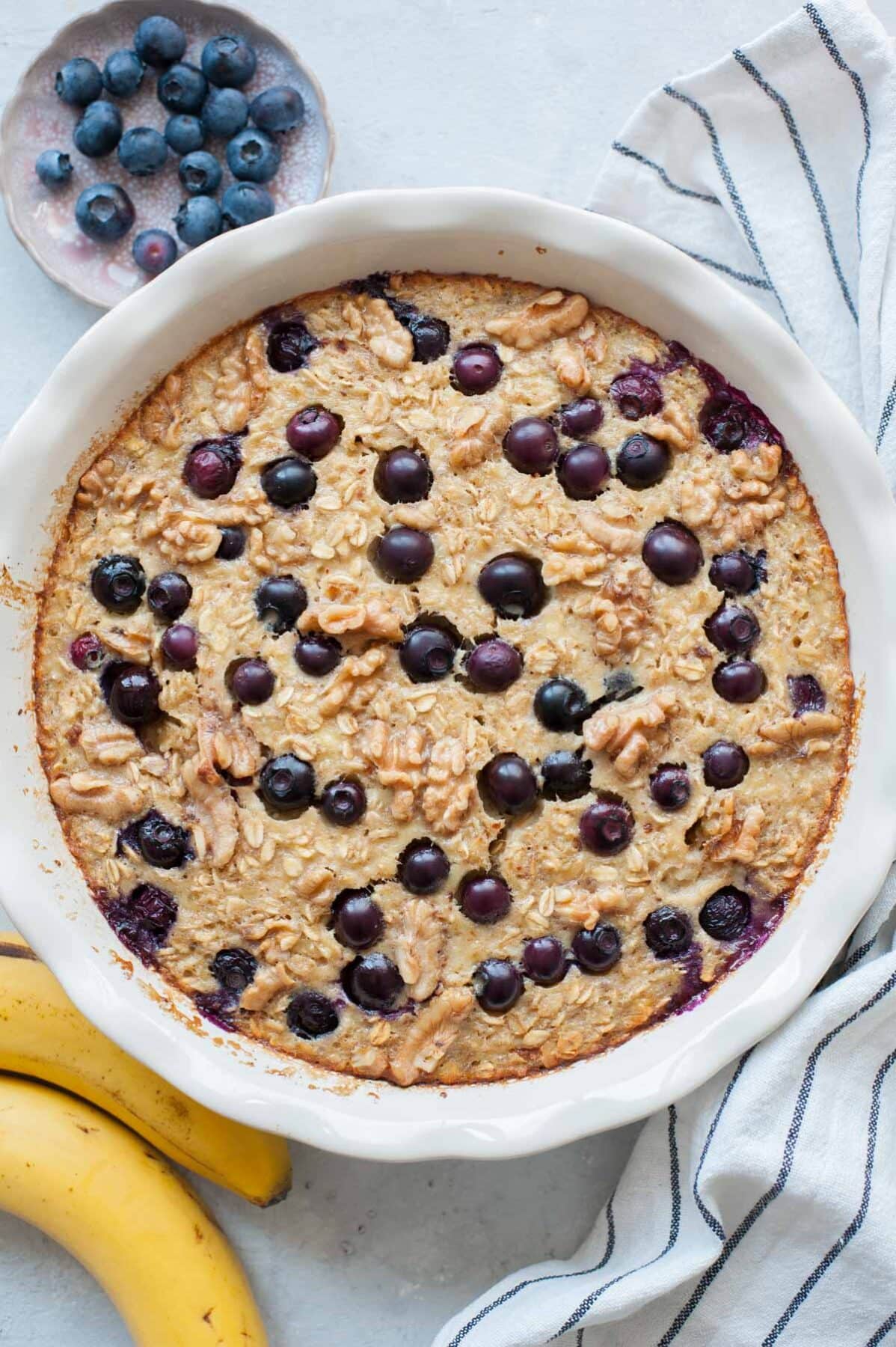 Baked oatmeal on a white table with striped towel near a banana and plate of blueberries