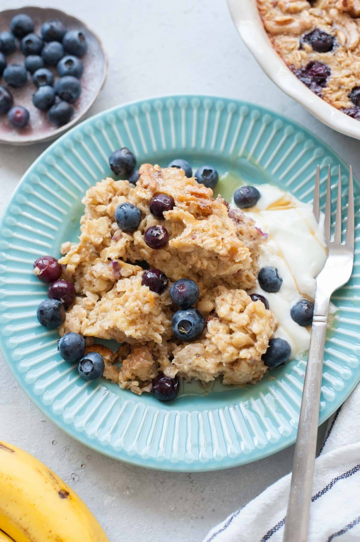 Baked oatmeal with blueberries and banana on a light blue plate, served with yogurt and maple syrup.