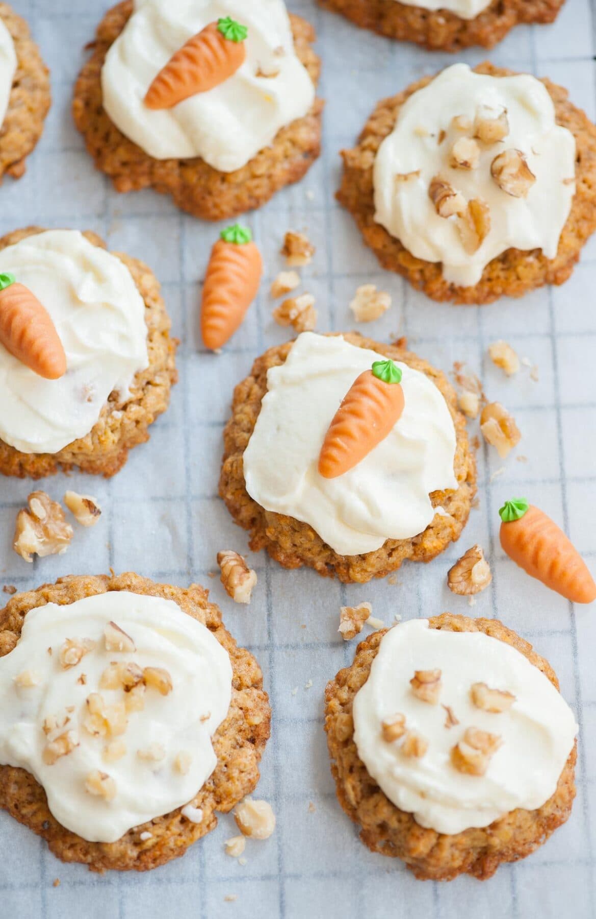 Carrot cake cookies with cream cheese frosting on a piece of parchment paper, topped with marzipan carrots and walnuts.