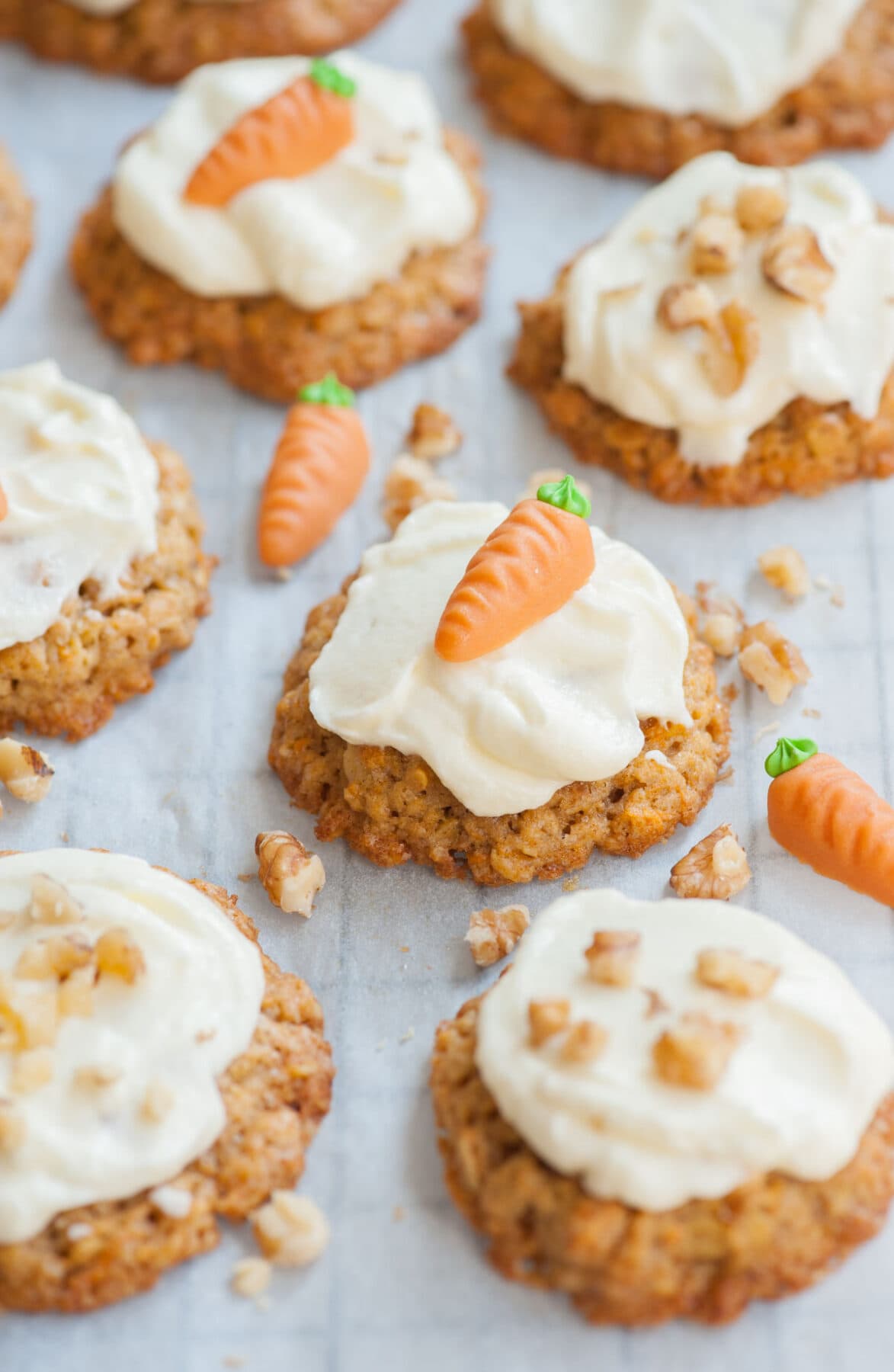 Carrot cake cookies with cream cheese frosting on a piece of parchment paper, topped with marzipan carrots and walnuts.