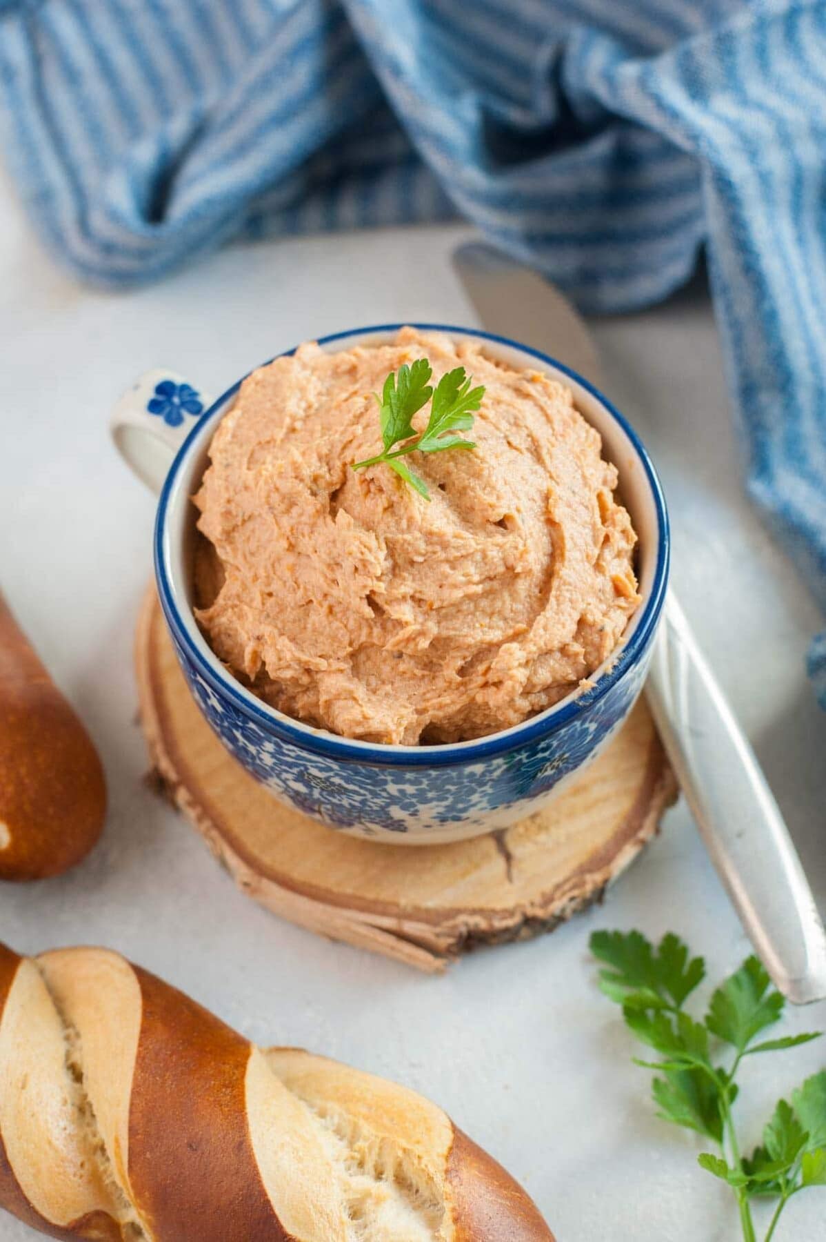 Chicken spread in a blue cup topped with parsley. Kitchen cloth and pretzels in the background.