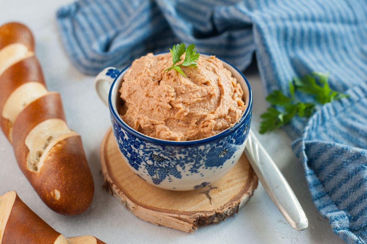 Chicken spread in a blue cup topped with parsley. Kitchen cloth and pretzels in the background.