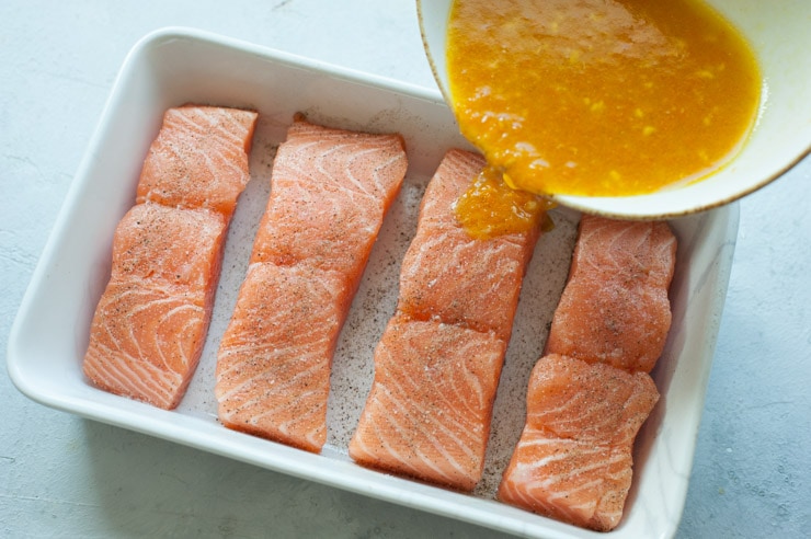 Orange maple sauce is being poured over salmon fillets in a white baking dish.