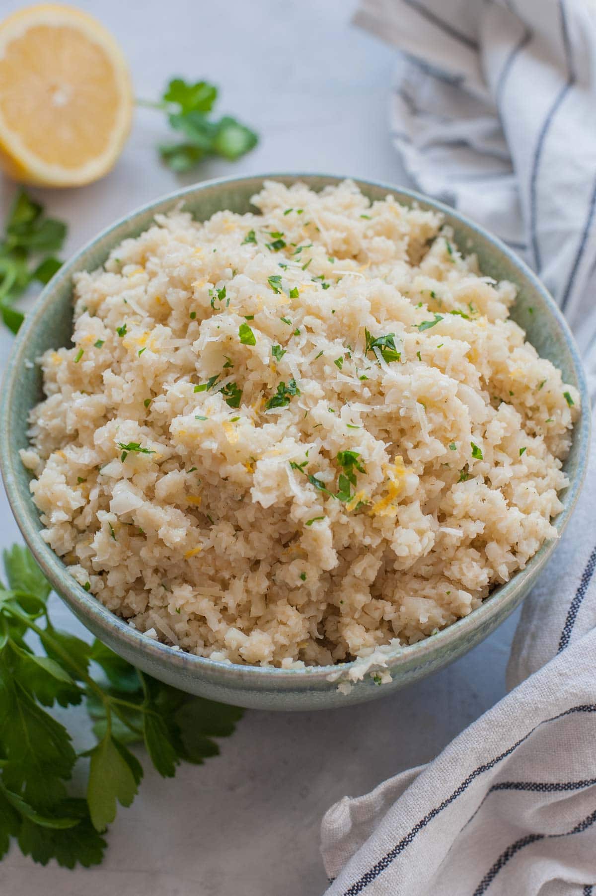 Parmesan cauliflower rice in a green bowl. Kitchen cloth and parsley leaved in the background.