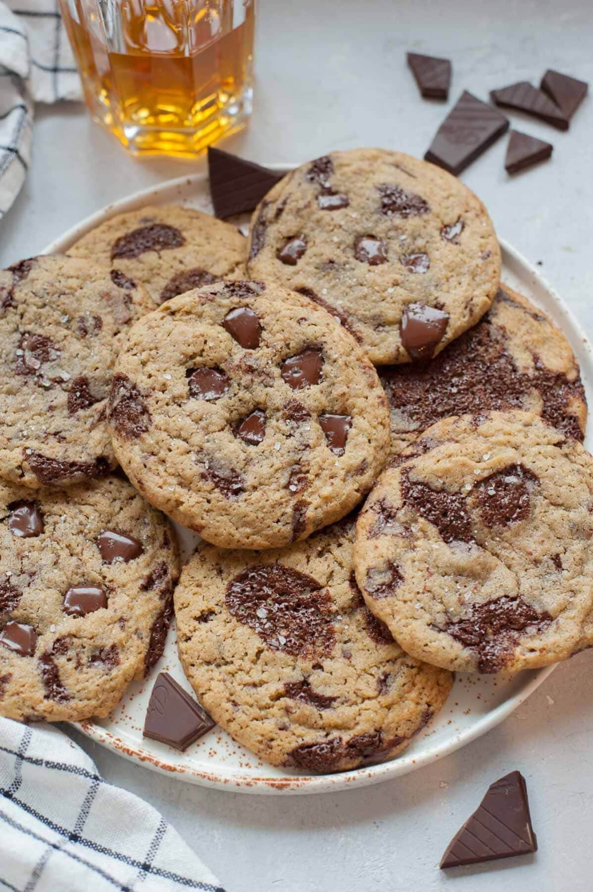 Rye chocolate chip cookies on a white plate. A glass with bourbon and chopped chocolate in the background.