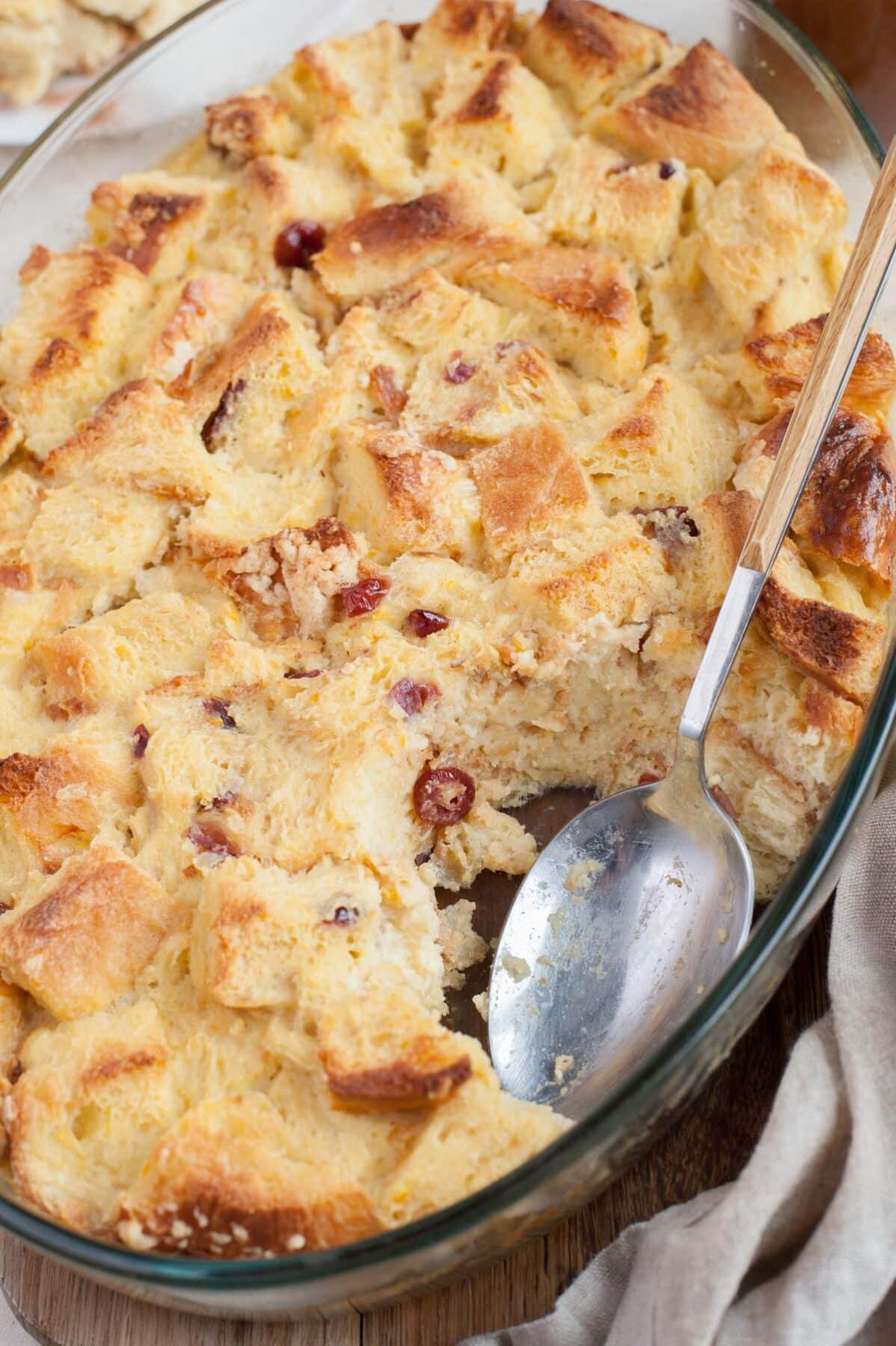 A close up photo of brioche bread pudding in a baking dish with a serving missing. A large spoon on the side.