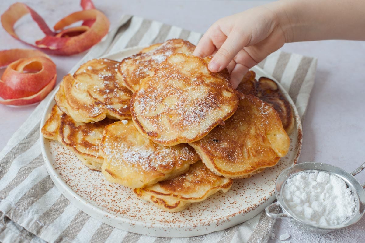 Apple pancakes on a white plate. One pancakes is being held by a child's hand.