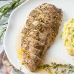 Baked turkey tenderloin on a white plate. Mashed potatoes and fresh herbs in the background.