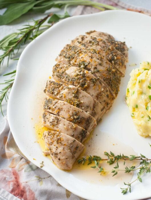 Baked turkey tenderloin on a white plate. Mashed potatoes and fresh herbs in the background.