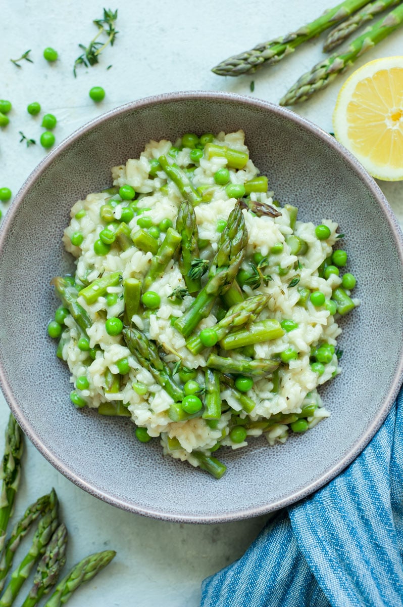 Asparagus risotto with peas in a violet bowl with asparagus and lemons in the background.