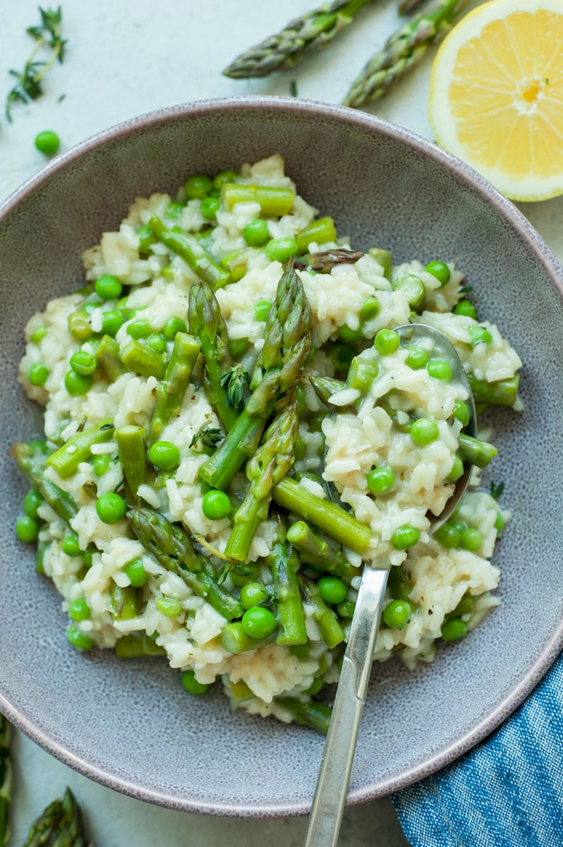 Asparagus risotto with peas in a violet bowl with asparagus and lemons in the background.