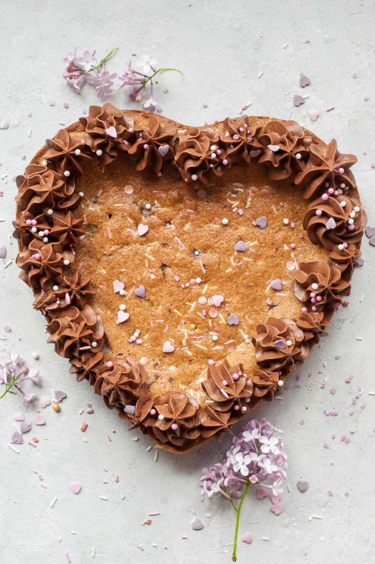 Chocolate chip cookie cake with chocolate frosting on a grey background. Sprinkles and flowers scattered around.